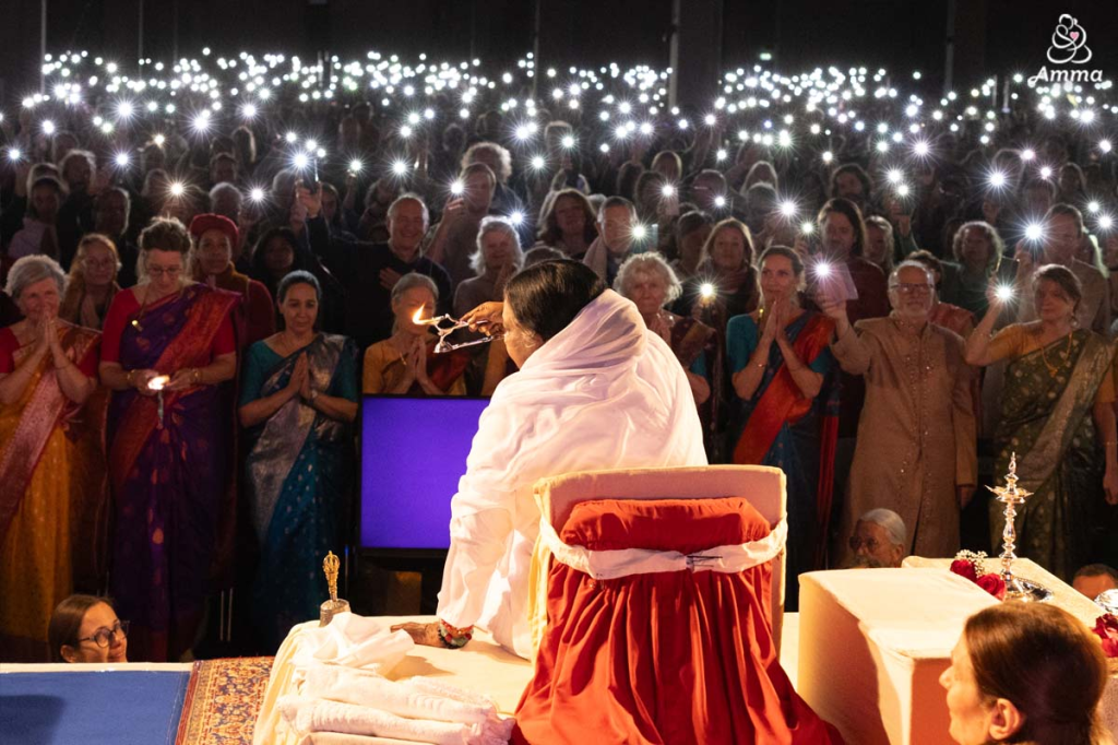 Amma lighting an oil lamp while addressing a large audience in Rijswijk, Netherlands, with attendees holding up mobile phones that illuminate the hall like stars.