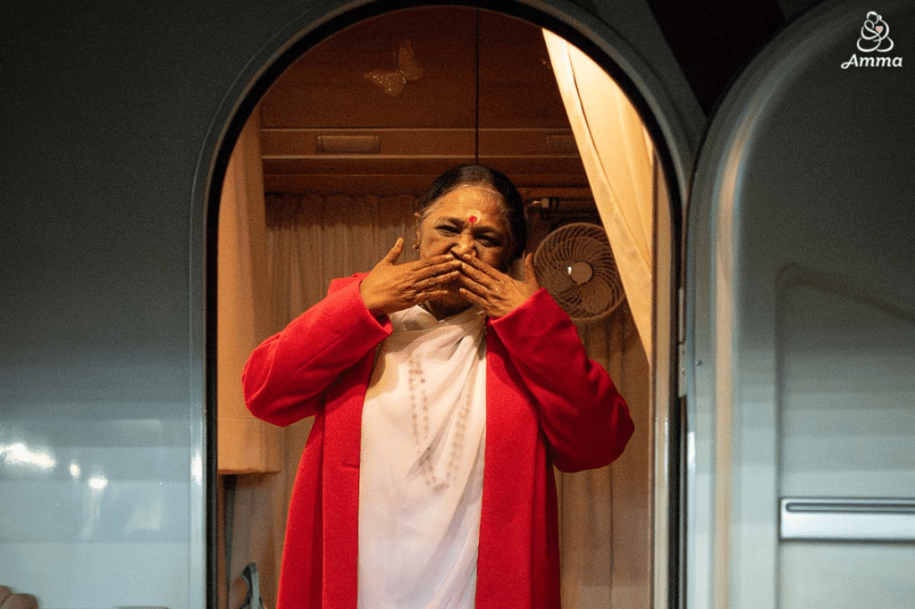 Amma blowing kisses while standing at the doorway of her vehicle, wearing a red robe, expressing love to her devotees.